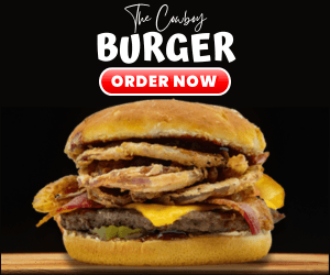 A close-up of 'The Cowboy Burger' featuring a thick patty topped with BBQ sauce, bacon, onion strings, and melted cheese on a toasted bun, set against a dark background.