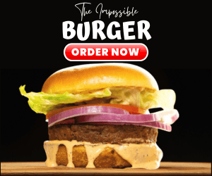 A close-up of The Impossible Burger, featuring a plant-based patty, lettuce, tomato, red onion, and special sauce, on a toasted bun with a black background.