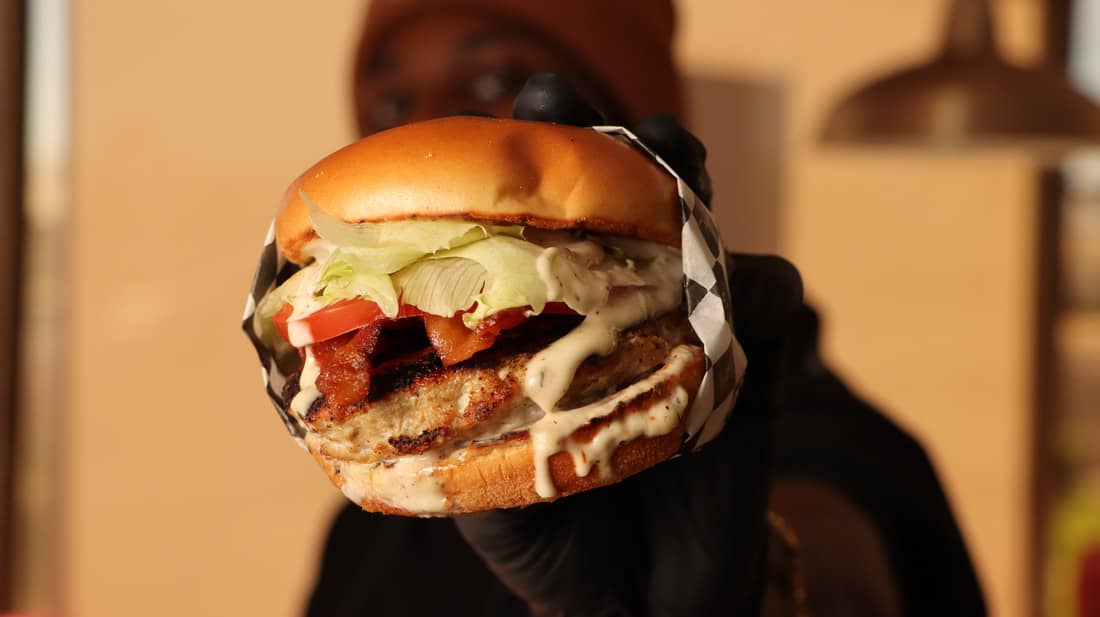 A person wearing black gloves holds a large burger with lettuce, tomato, bacon, and sauce, against a blurred background.